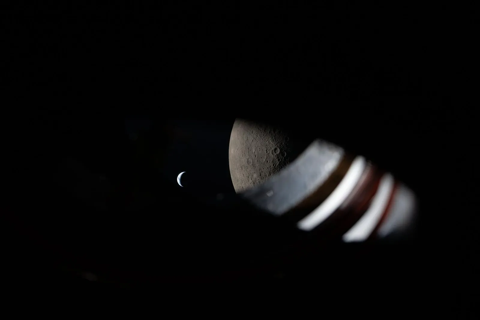 Through the edge of an Orion window, a thin crescent Earth hangs in darkness next to the half-lit, cratered Moon