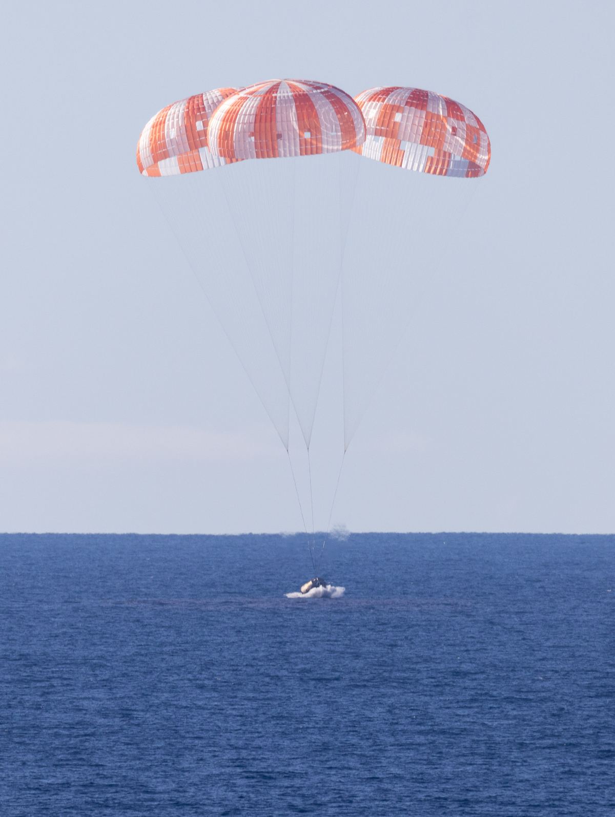 Orion under three orange and white parachutes just above the Pacific Ocean