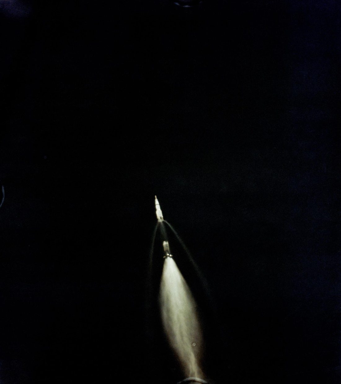 Apollo 11's upper stages pulling away from the spent first stage, with a wide, pale exhaust plume against a black sky
