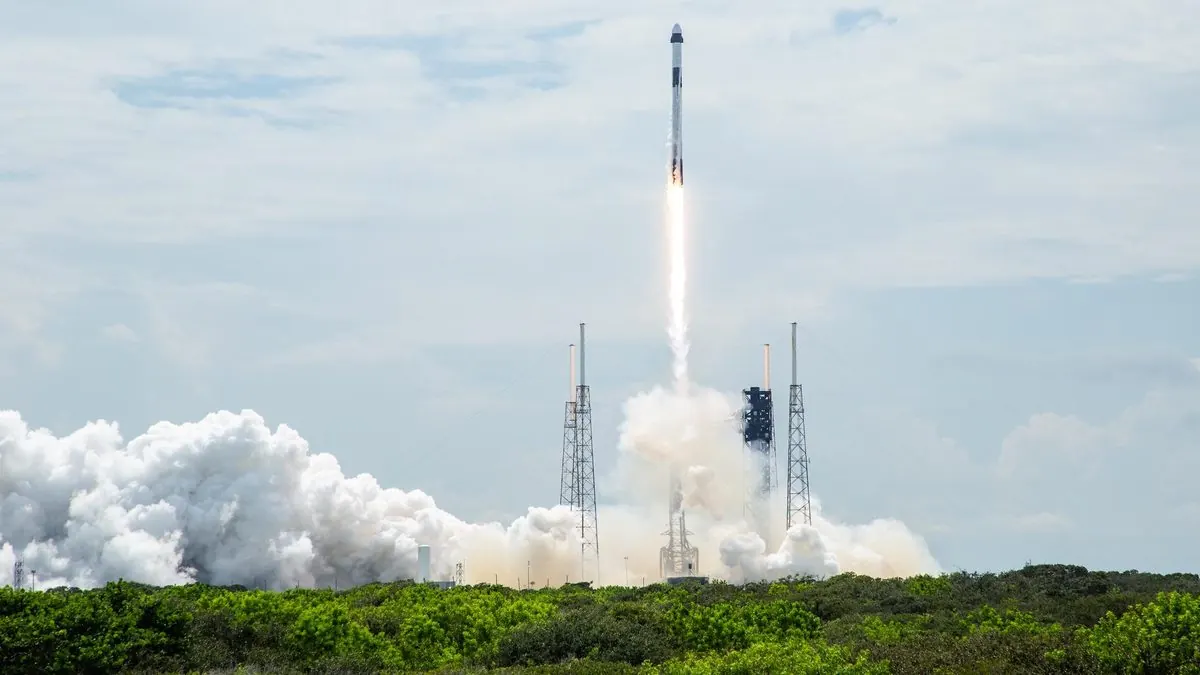 A Falcon 9 rocket climbing from its launch pad in daylight on a long bright flame