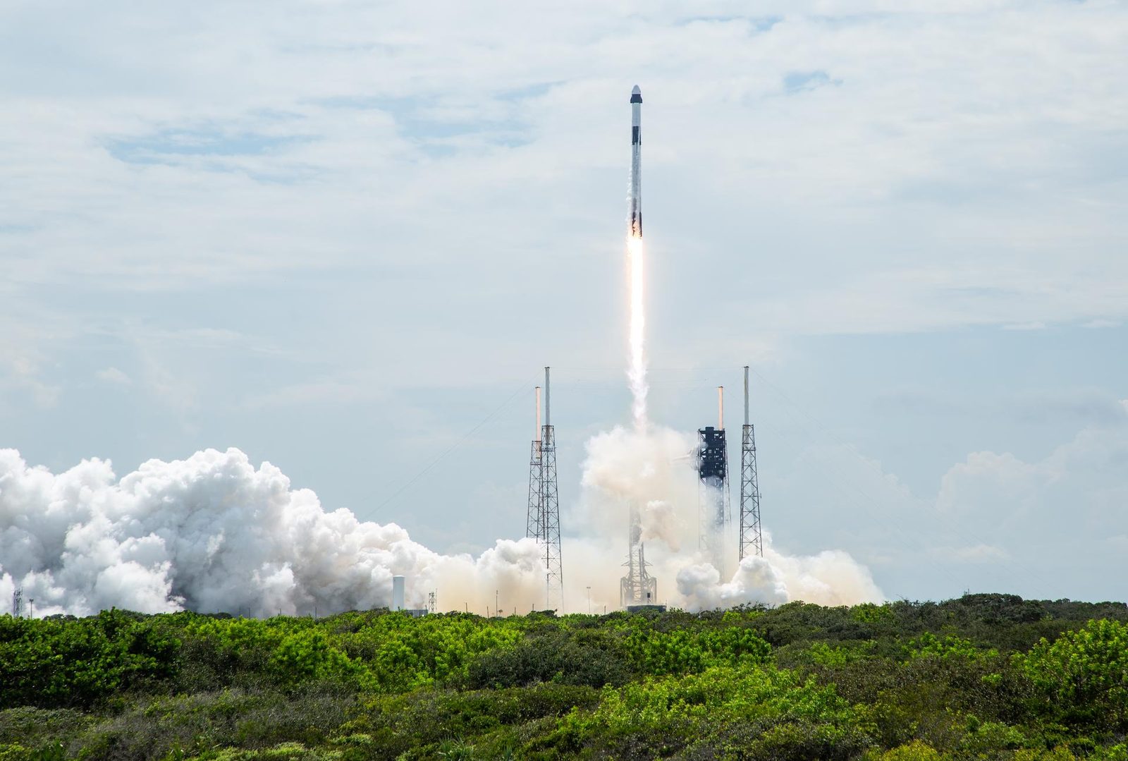 A Falcon 9 rocket climbing from its launch pad in daylight on a long bright flame, above clouds of white steam