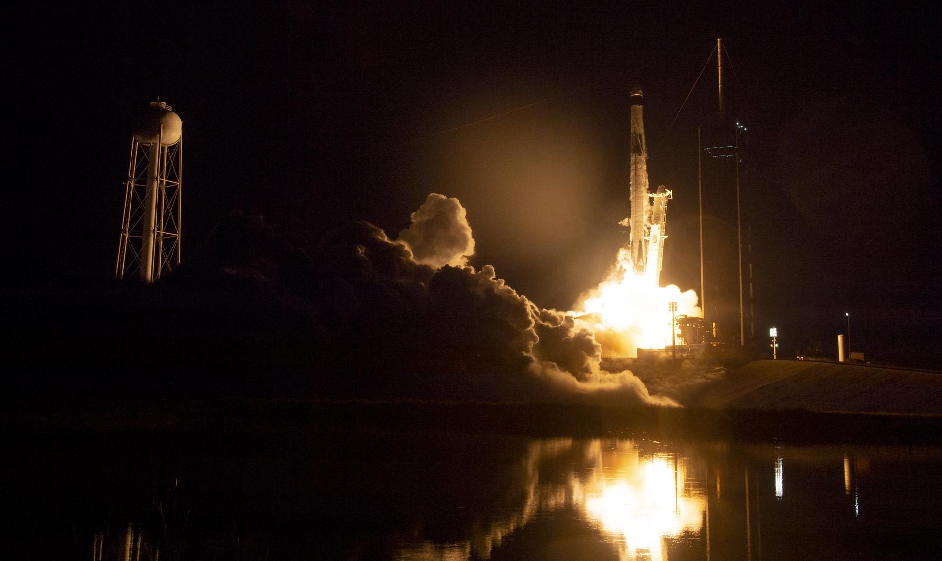 A Falcon 9 rocket lifts off at night on a bright column of flame, reflected in the water in front of the pad