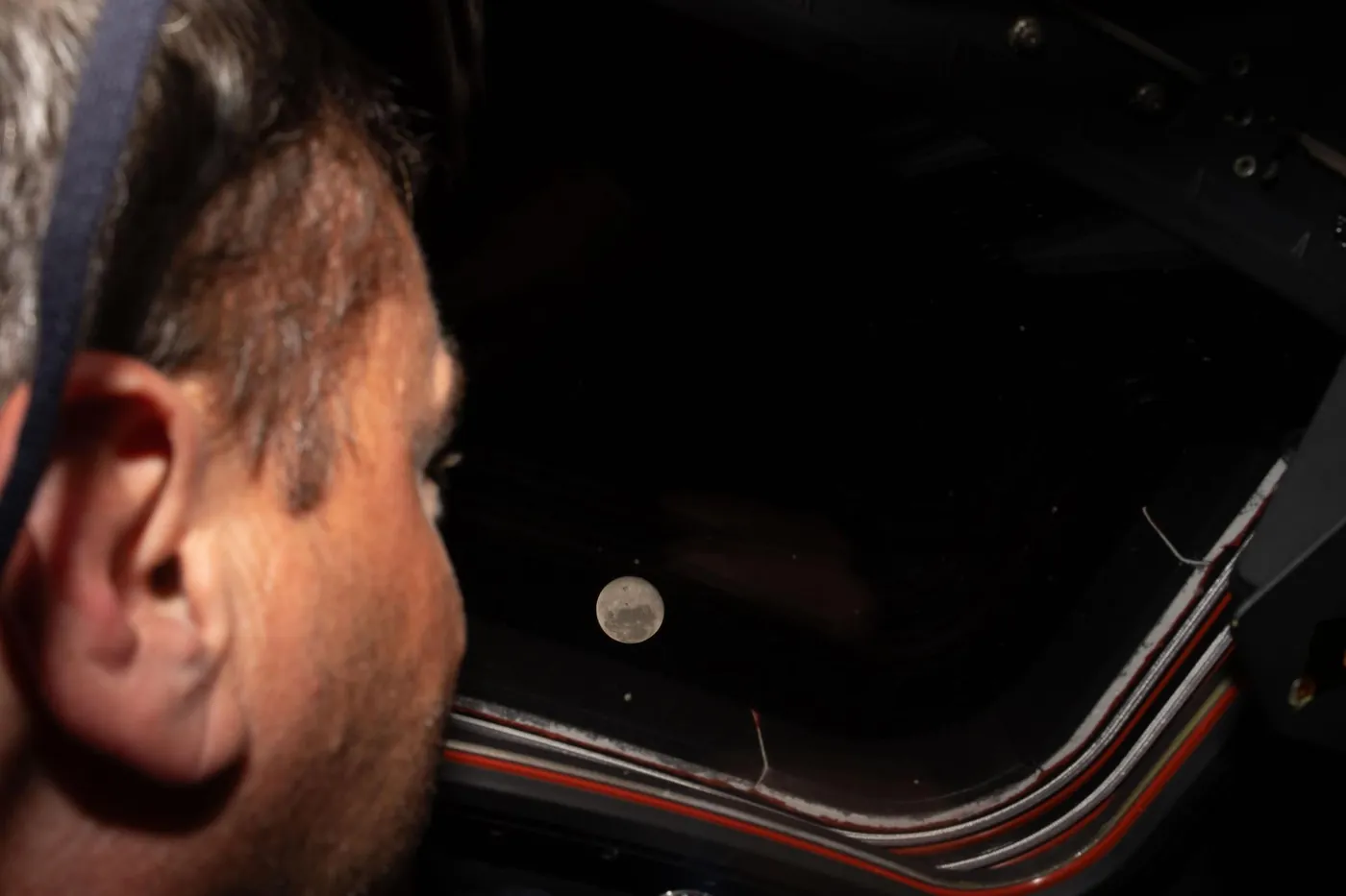 Artemis II commander Reid Wiseman, seen from behind, looking out of an Orion window at a small, bright Moon