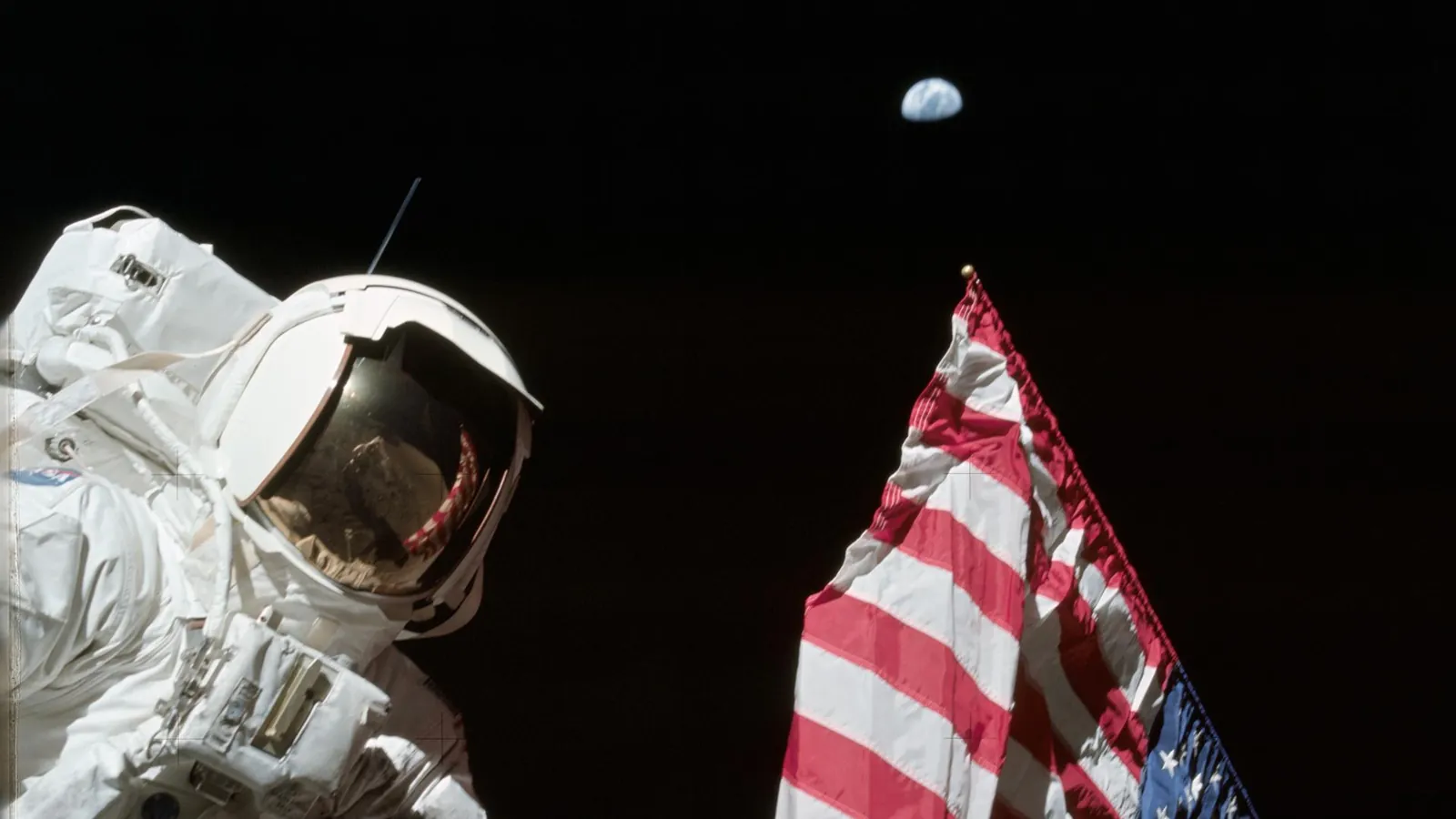 Apollo 17 astronaut Harrison Schmitt in a white spacesuit beside the US flag on the Moon, with a small Earth in the black sky above